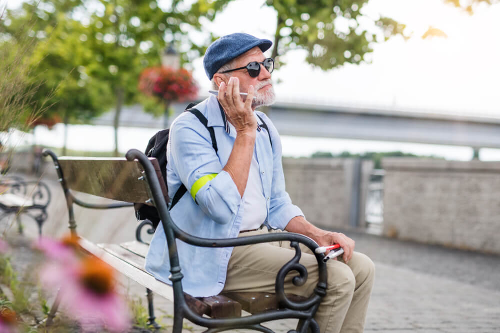 Foto de um senhor cego sentado em um banco de praça ouvindo algo em seu smartphone