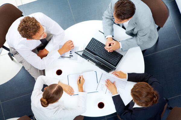 Foto vista de cima de quatro pessoas vestindo-se socialmente e sentadas em uma mesa redonda discutindo sobre trabalho